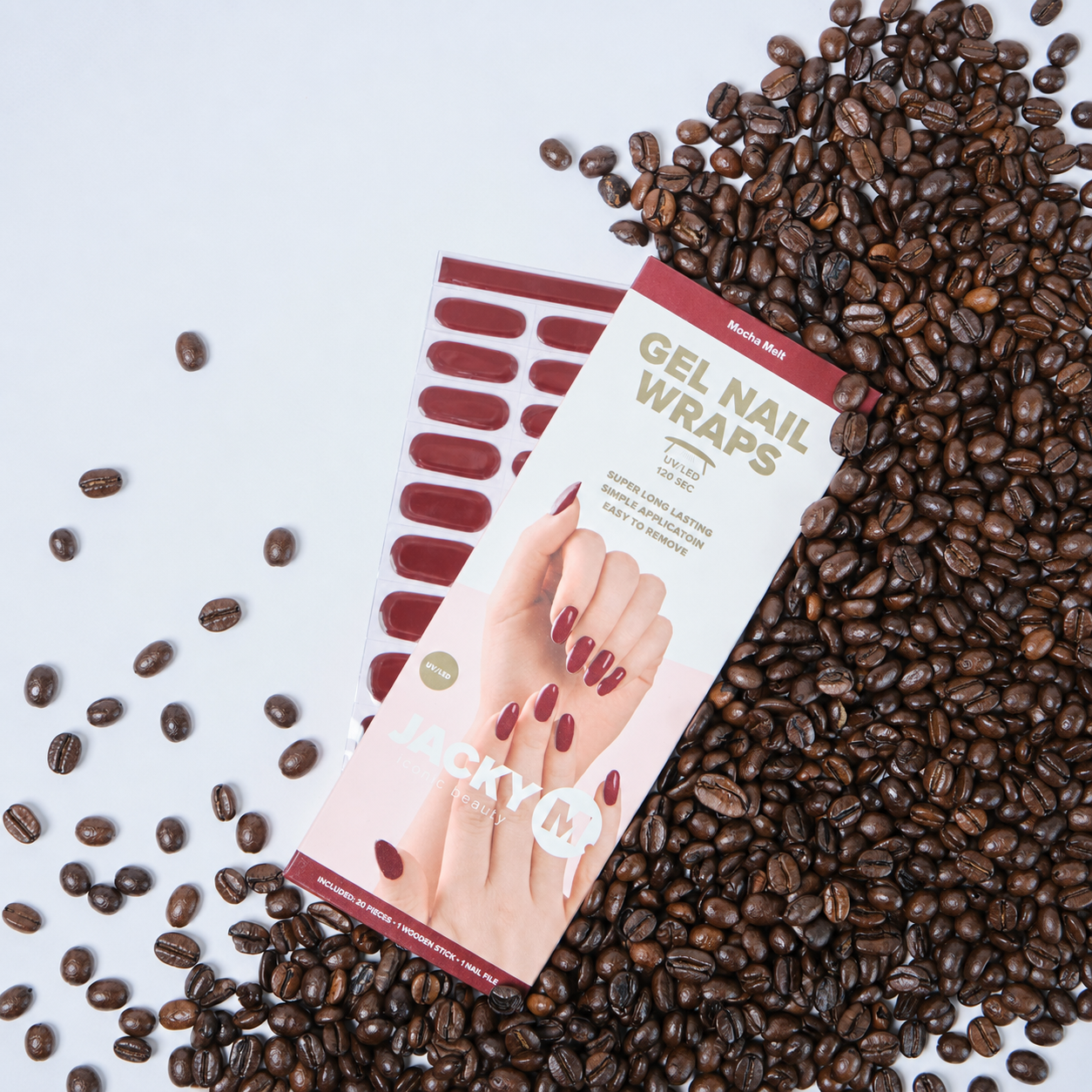 A package of Jacky M Gel Nail Wraps in the shade 'Ruby Pink', displayed diagonally on top of a sheet of matching nail wraps. The package is styled on a white background and surrounded by scattered dark brown coffee beans.